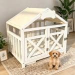 A detailed view of a white dog villa crate with a bone emblem above open doors, showing a thick plush white bed and a cream canvas roof.