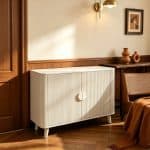 A modern cream-colored fluted accent cabinet with distinctive curved edges and sleek half-moon handles, styled with natural woven baskets and modern wall art in a sunlit room.